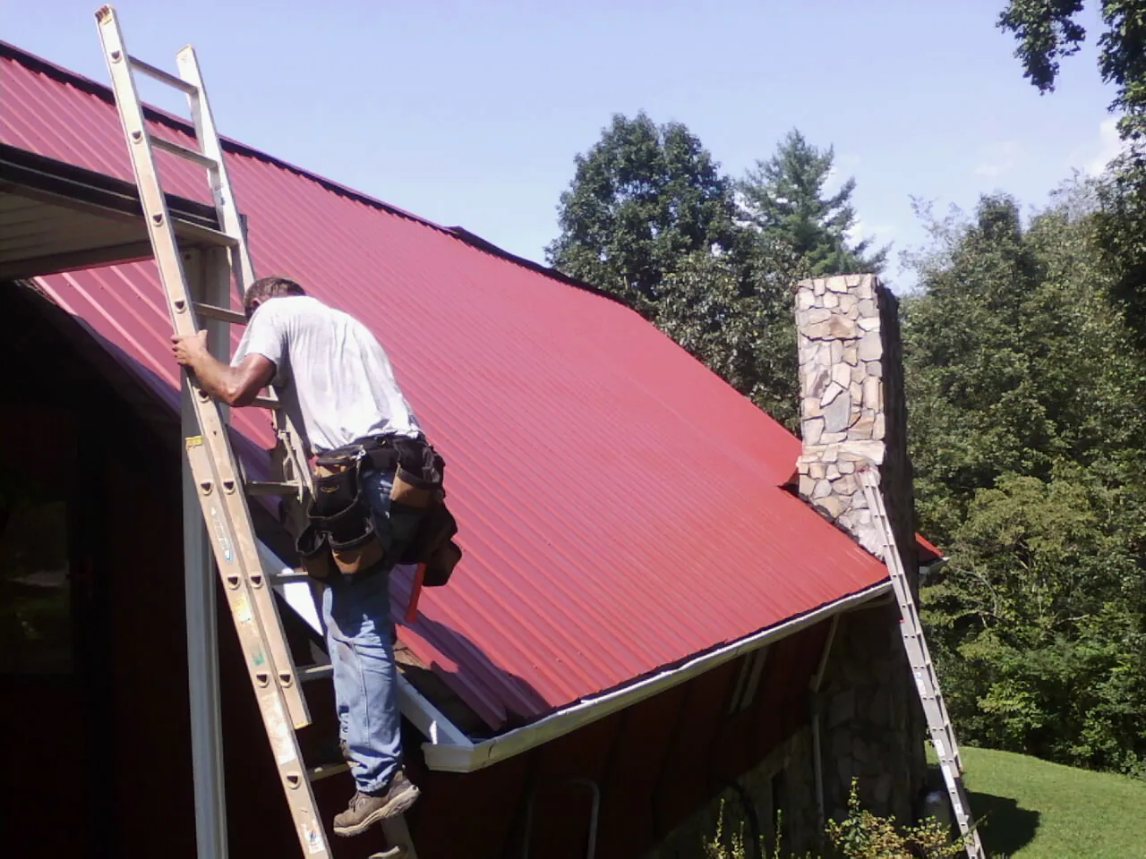 Roofer on a ladder beside a red metal roof and stone chimney — Soffit & Fascia Repair by Roofing Brothers in Los Alamos
